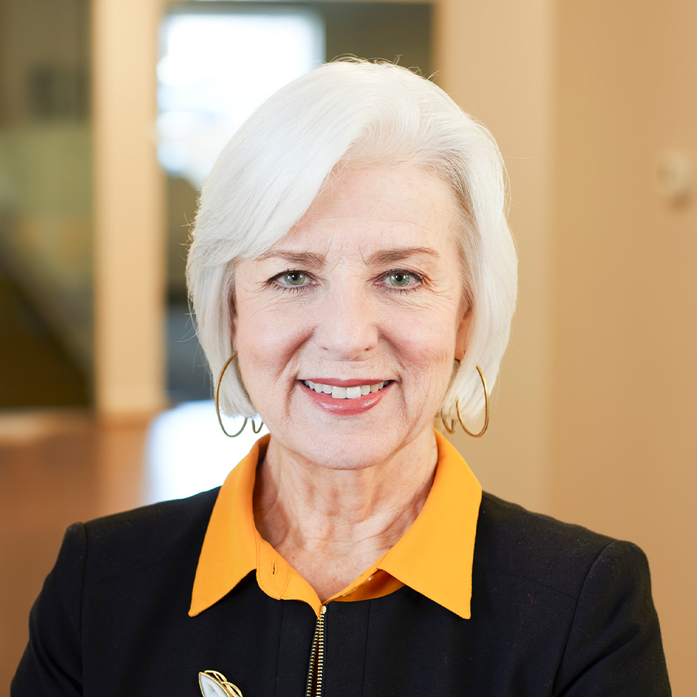 Cynthia, a woman with white hair, a yellow shirt, and a black jacket, smiles at the camera.