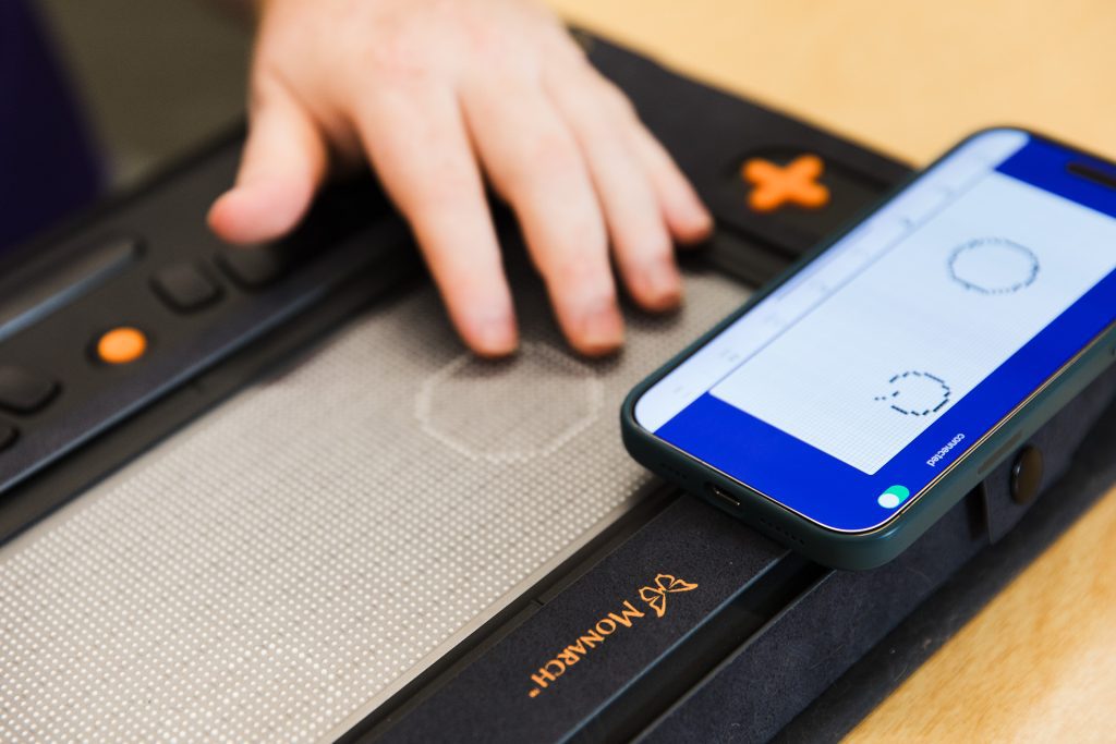 A phone rests on the Monarch with the Wing It app open, showing two drawings. On the Monarch screen below, we see the two shapes in tactile form.