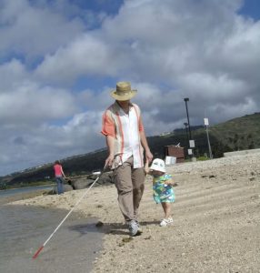Tim walks along the beach with his cane in his right hand, holding his daughters hand on his left as she walks beside him/