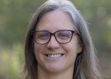 Kirsten stands outside with the background blurred, smiling at the camera. She has light silver hair and black glasses.