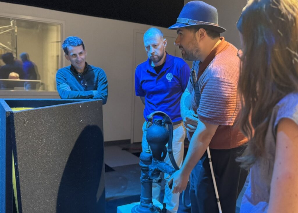 Four adults, one with a cane, stand around the 3-D water pump and feel the exhibit.