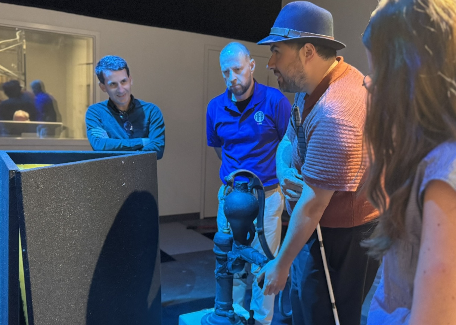 Four adults, one with a cane, stand around the 3-D water pump and feel the exhibit.
