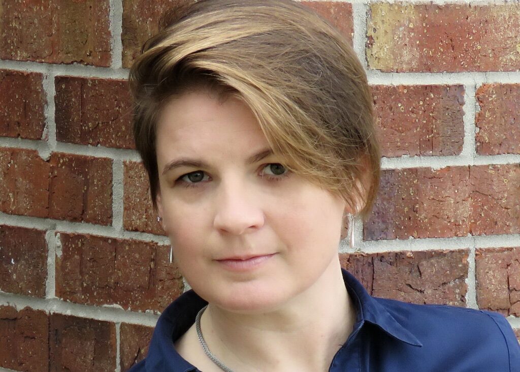 Jenn, a woman with short brown hair, stands smiling against a brick building.