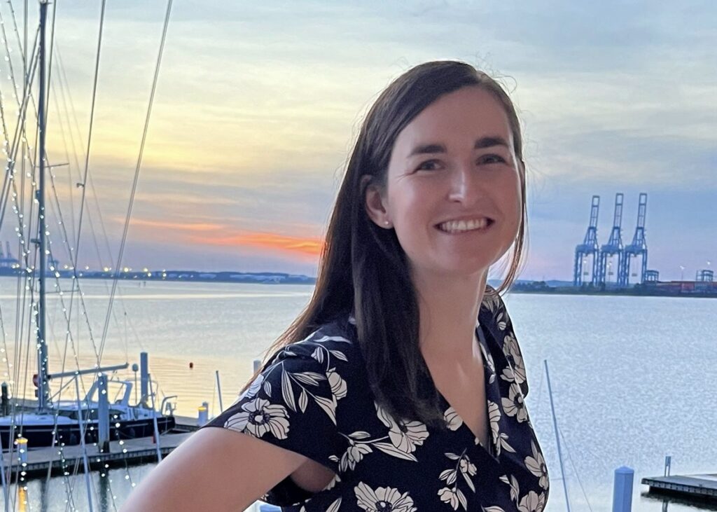 Katy, a woman with dark brown hair, stands and smiles in front of a sunset and what appears to be the ocean.