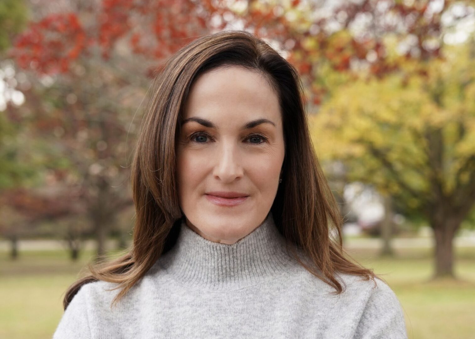Missy, a woman with brown hair and a grey sweater, stands in front of trees with red and yellow leaves.
