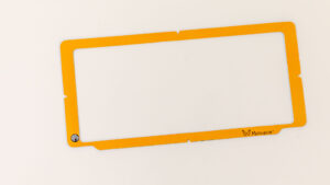 The orange plastic Monarch Masking Frame on a white background. In the bottom left corner is a small APH logo, and in the bottom right is the Monarch logo.