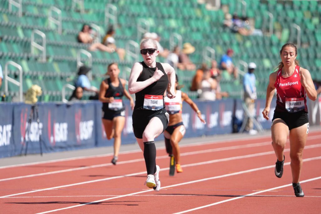 Addie, dressed in an all black running outfit with dark black sunglasses, runs across the track.