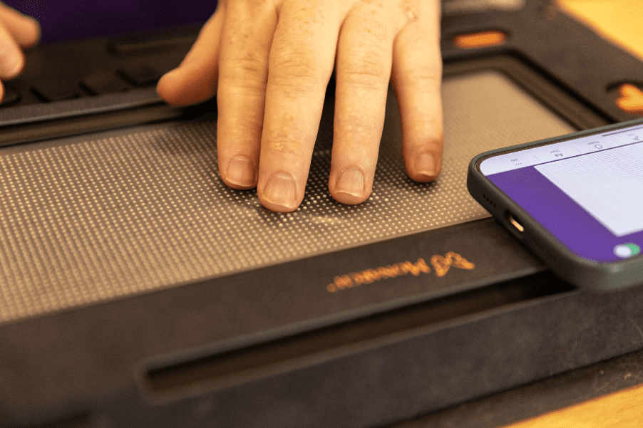 A hand traces a star shape on the Monarch display while a phone showing the WingIt app lays near by.