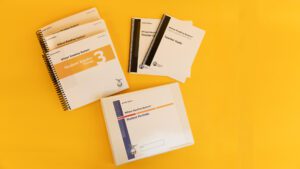 Wilson Reading System IV braille step three on a yellow background, including three volumes of the student reader 3, braille and large print versions of the teacher guide, and the braille student portfolio.