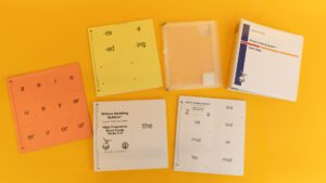 The card set components laid out on a yellow background, including a three-ring binder, binder inserts, and four sets of cards in a variety of colors with print and braille labels.