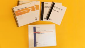 Wilson Reading System IV braille step two on a yellow background, including two volumes of the student reader 2, braille and large print versions of the teacher guide, and the braille student portfolio.