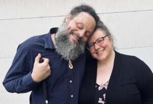 John, wearing a navy button down and holding his cane smiles while he leans into his wife Anni.