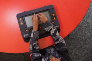 A student reaches his arms out towards the Monarch. He is sitting at a bright red classroom table, the Monarch is resting on the table.
