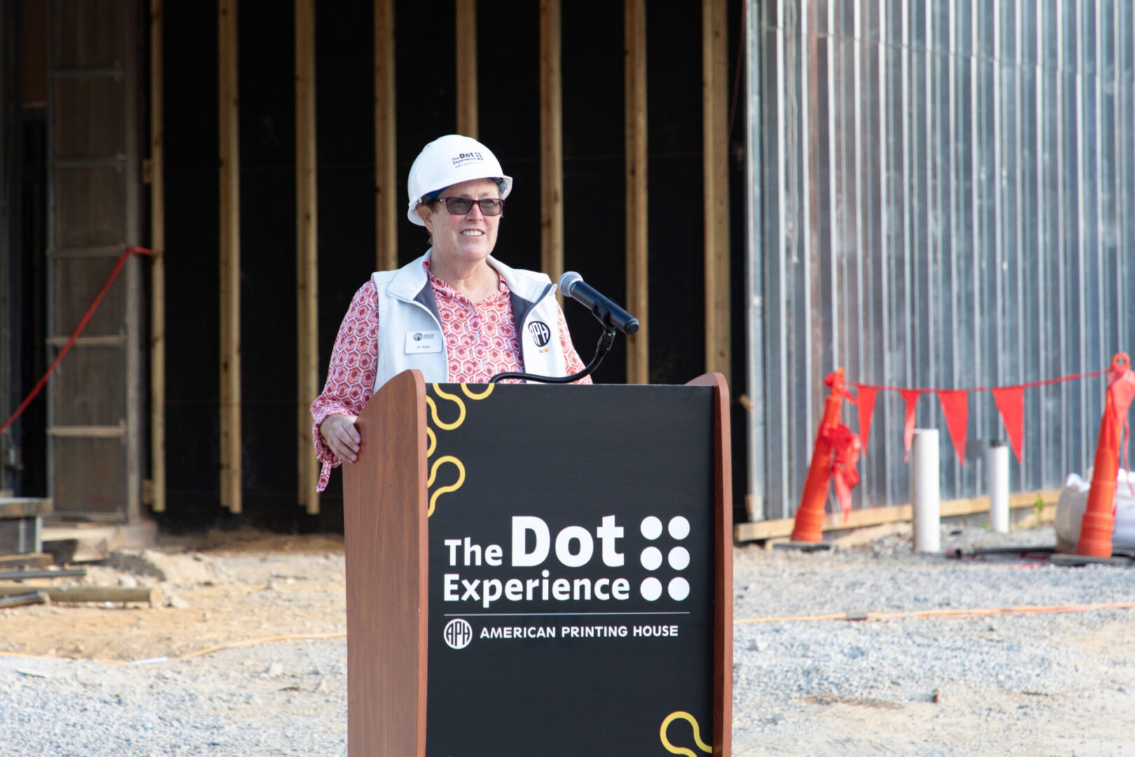 Jo Haas stands behind a podium that says "The Dot Experience" while wearing a white hard hat, also reading "The Dot Experience."