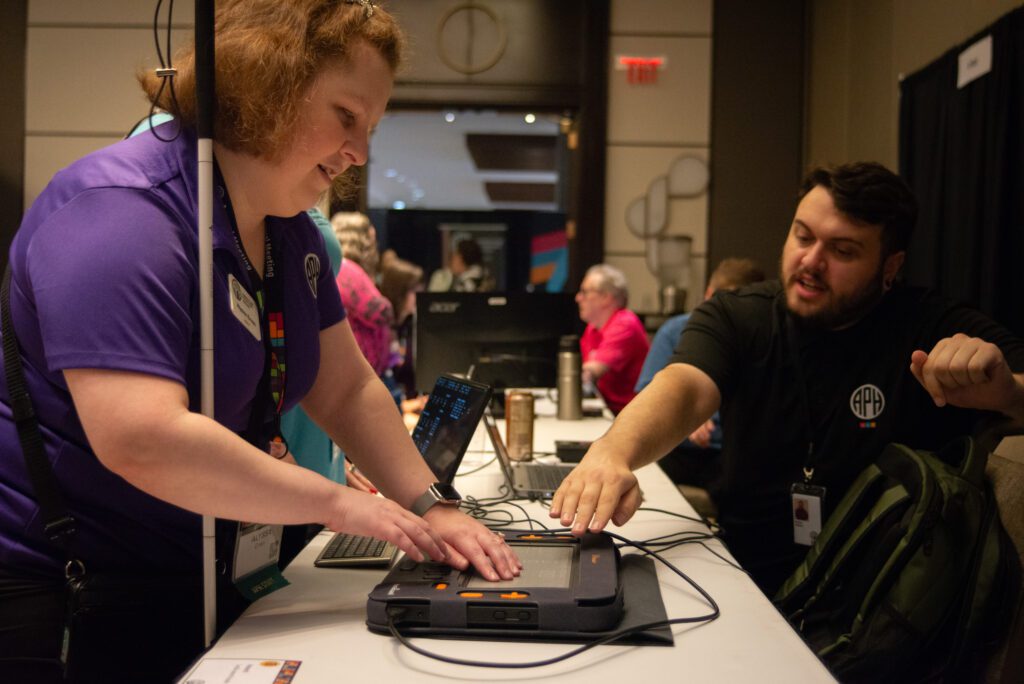 A Monarch sits on a table. A woman holding a cane is reaching down to read what is on the display, while a man sitting behind the table leans forward to guide her around the device.