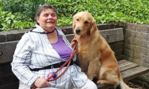 Debee sitting on a wooden bench outdoors, holding the leash of a golden retriever dog seated next to her. Debbi is wearing a striped long-sleeve shirt over a purple top and white pants. She has short hair and is smiling gently. The background features green leafy plants and a brick wall. The dog is looking forward with a calm expression, sitting close to Debee. The setting appears peaceful and natural.