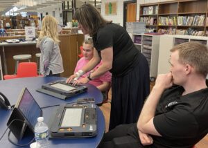 A classroom table shows a student feeling the Monarch with a teacher. Next to them is a man in an APH polo with a Monarch, connected a computer screen.