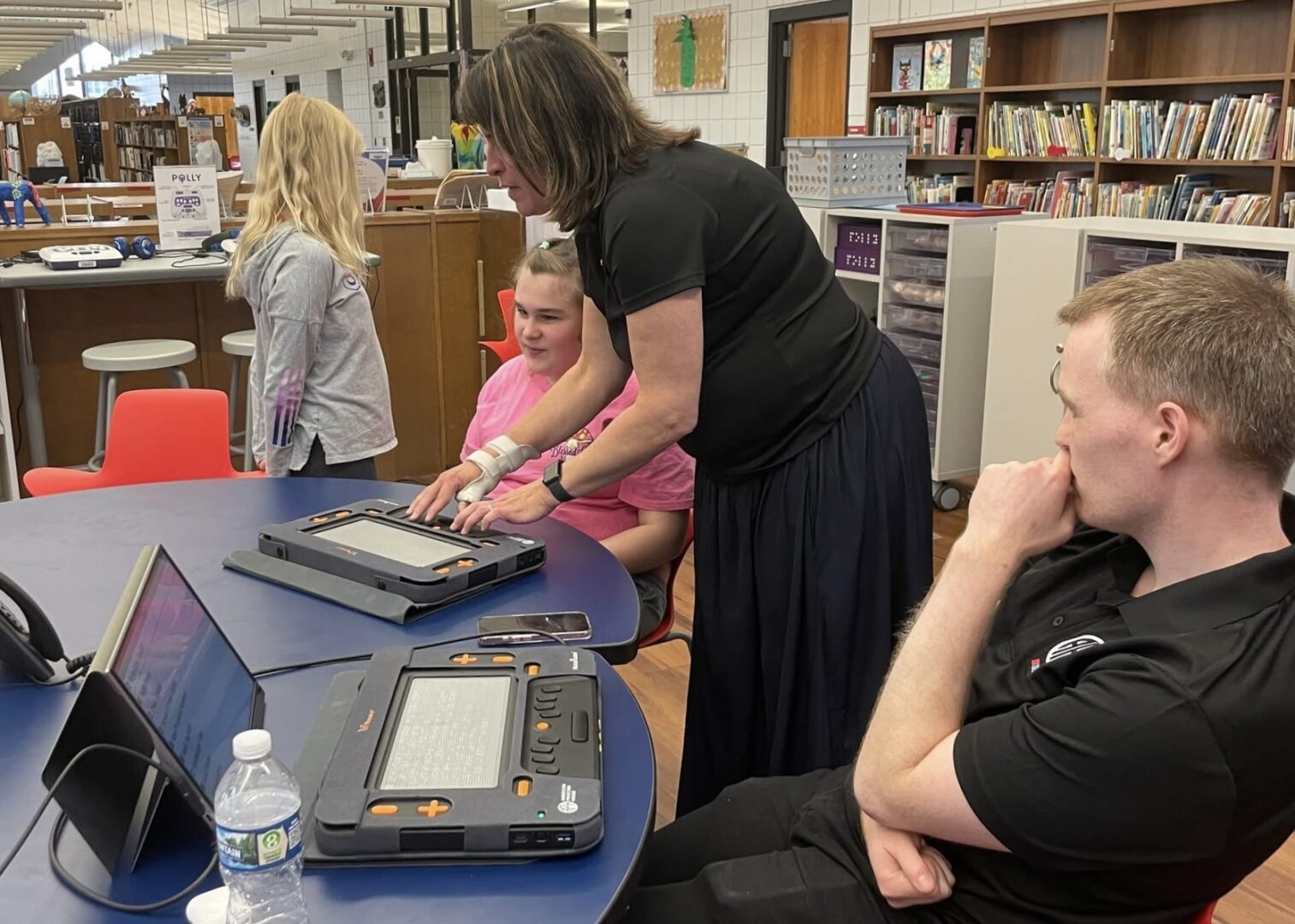 A classroom table shows a student feeling the Monarch with a teacher. Next to them is a man in an APH polo with a Monarch, connected a computer screen.