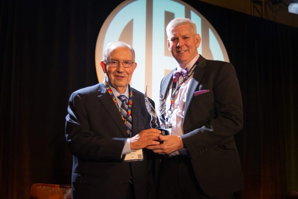 Dr. Wiener stands next to Dr. Meador, both holding the Wings of Freedom Award as they smile at the camera. Behind them a large image of the APH logo can be seen.