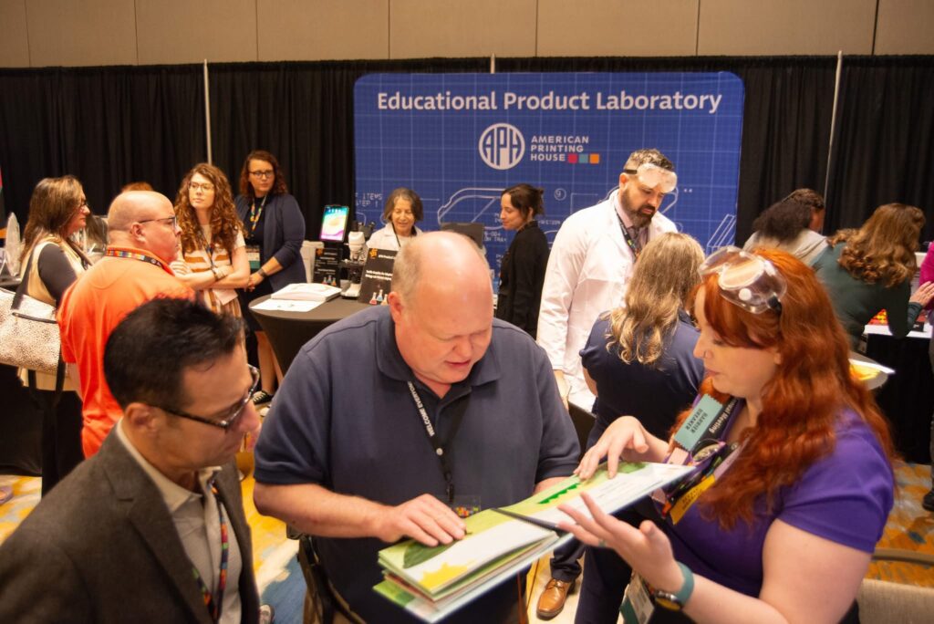 Three adults look closely at a tactile book. Behind them a large crowd of people moving between booths of products can be seen.