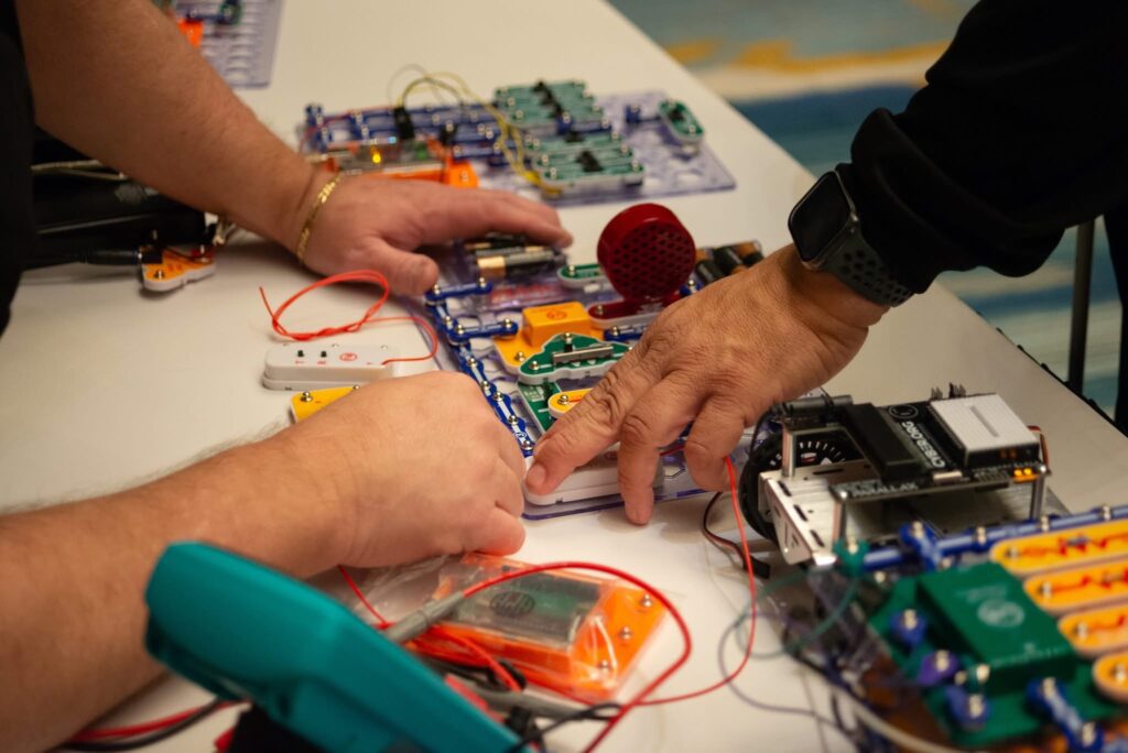 An attendees hand rests on top of the snap circuits product, where you can see another hand being used to guide and instruct on the product.