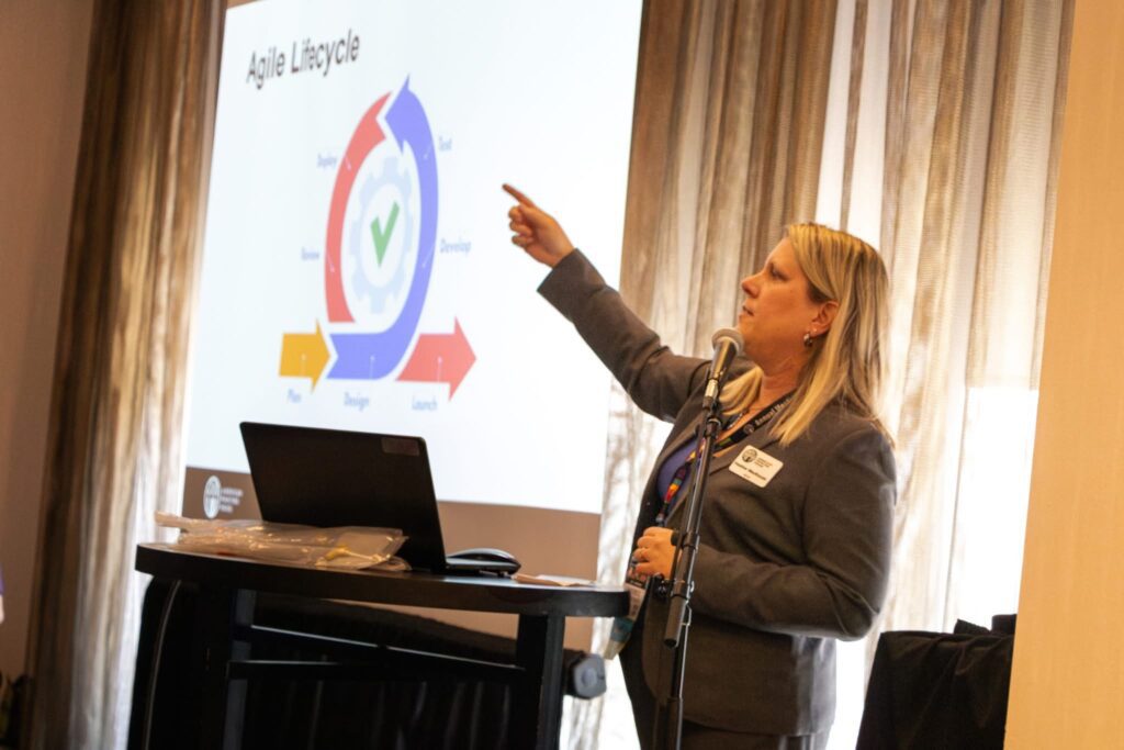 Heather Spence, APH employee, stands in a grey suit speaking behind a podium. Using her right hand she points at a presentation behind her.