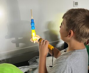 A student holds a figurine in front of a wall. In his other hand, he holds a flashlight to show the shadows of the figurine on the wall.