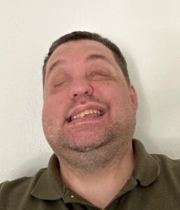 Liam has short dark hair and has a short beard, wearing a dark green collared shirt. He is facing the camera, with his eyes closed and smiling.