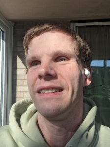 Michael with short light brown hair wearing a light green hoodie. He is smiling slightly and looking slightly to his left. He has wireless earbuds in his ears. The background includes a window with sunlight illuminating part of his face and a brick wall.