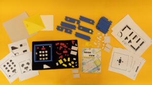 All of the components of the Room With a View kit including tactile graphics, blue foam building block walls, a variety of 3D pieces, and a print guidebook all laid out on a yellow background.