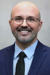 Ben a smiling, he has a bald head and a neatly trimmed beard and mustache. He is wearing glasses, a light-blue dress shirt, a black tie, and a dark blue suit jacket.