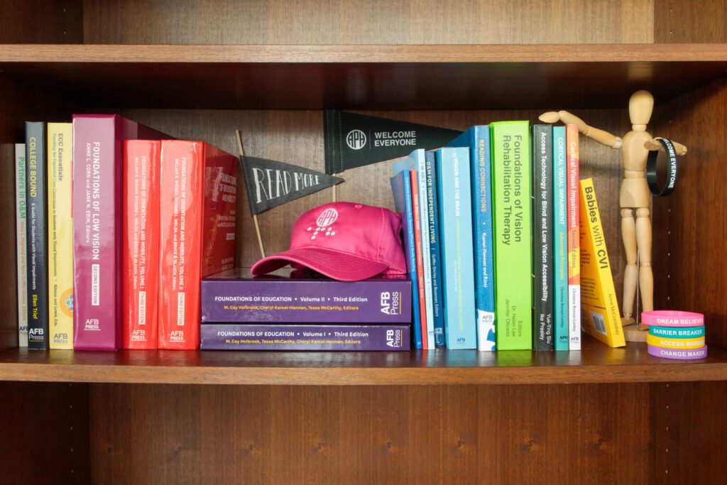 On a wooden shelf colorful volumes of APH press books can be seen stacked together. In the middle is a pink hat with the APH logo printed on the front.