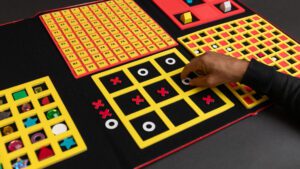 A hand placing a white circle in a yellow three-by-three grid in a game of tic tac toe. The grid is attached to a felt board that contains a variety of other grids used for different activities.
