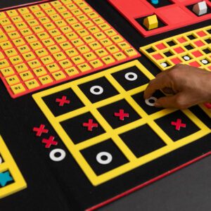 A hand placing a white circle in a yellow three-by-three grid in a game of tic tac toe. The grid is attached to a felt board that contains a variety of other grids used for different activities.