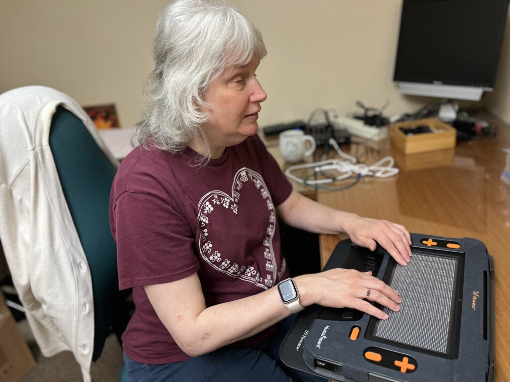 Chris, a woman who is blind, sits at her classroom desk. She has her hands on her Monarch, which is resting on her desk, while she plays a sudoku puzzle.