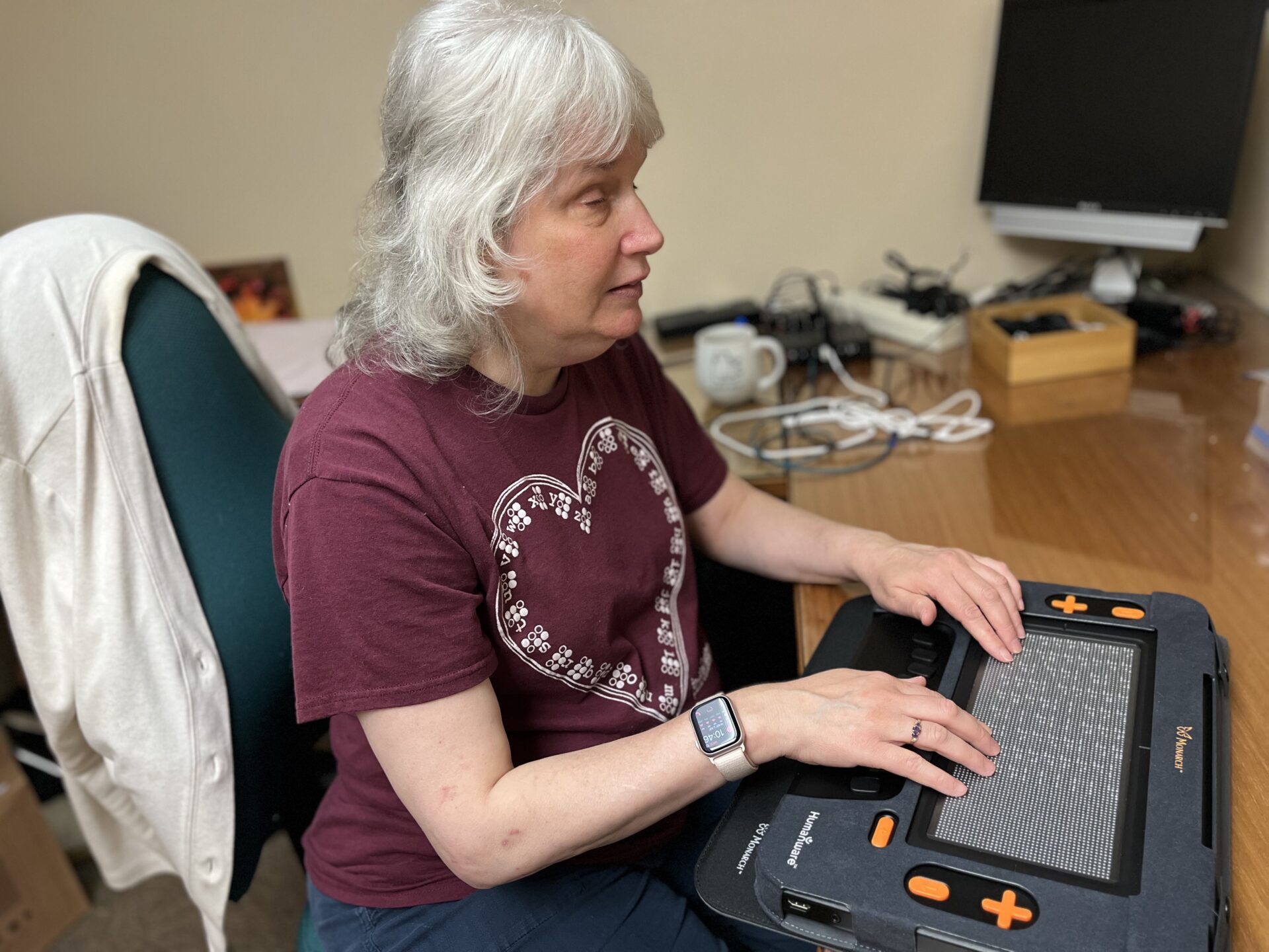 Chris, a woman who is blind, sits at her classroom desk. She has her hands on her Monarch, which is resting on her desk, while she plays a sudoku puzzle.