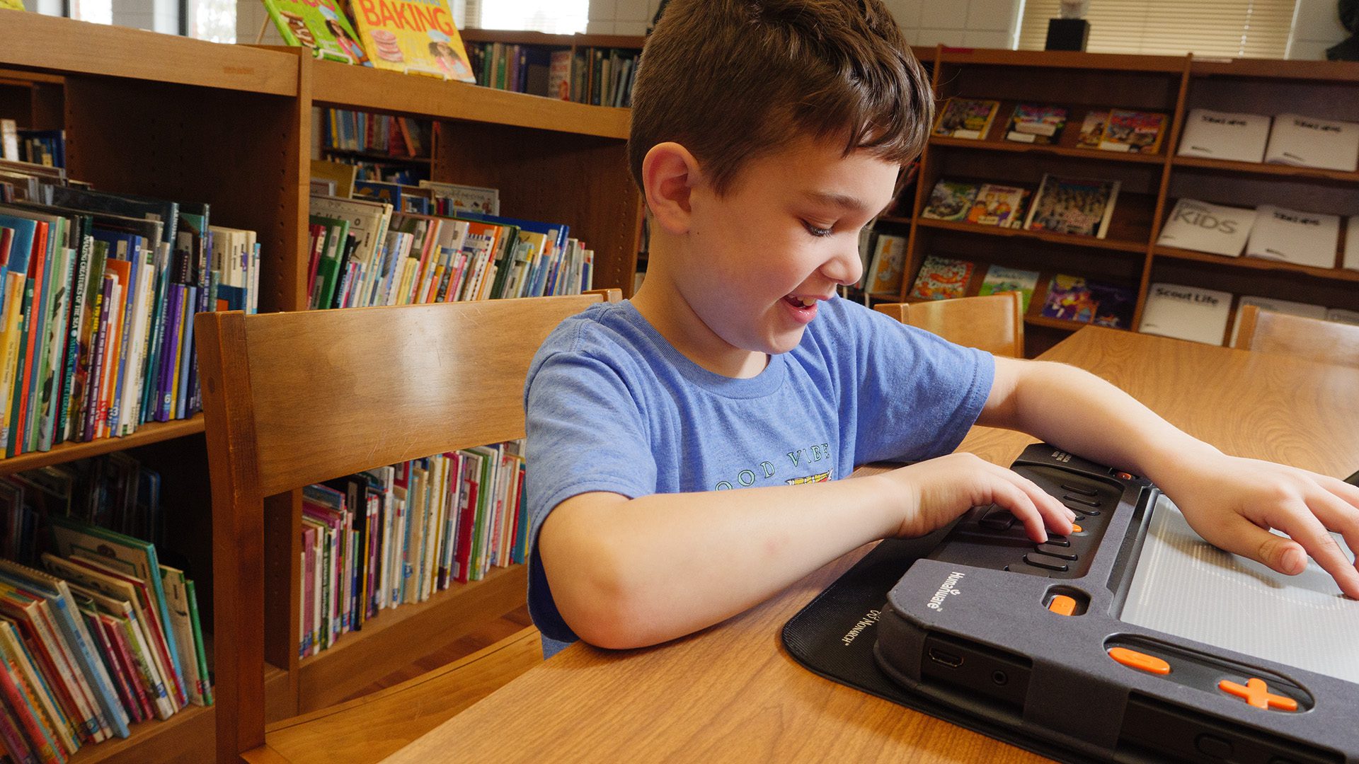A young boy sits at a table, with his hands on the Monarch, and smiles. Behind him are bookshelves.