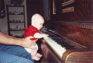 A young Patrick, around the age of 9 months, is being held up in front of the piano where he plays the keys.