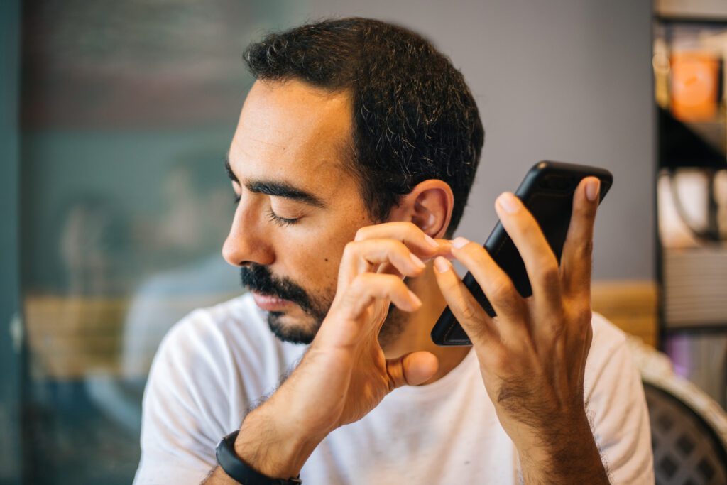A man holds his phone up towards his ear. His eyes are closed as he leans his ear towards the phone speaker.