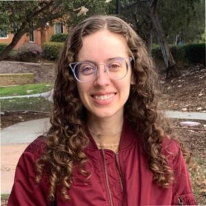 Isabela Figueira standing outside with trees in the background. She has long curly hair and wears glasses.