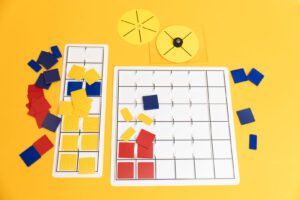 All the components of the MathBuilders Statistics kit laid out on a yellow background. There are two white graphing grid trays, two yellow spinners, and a variety of yellow, red, and blue tiles.