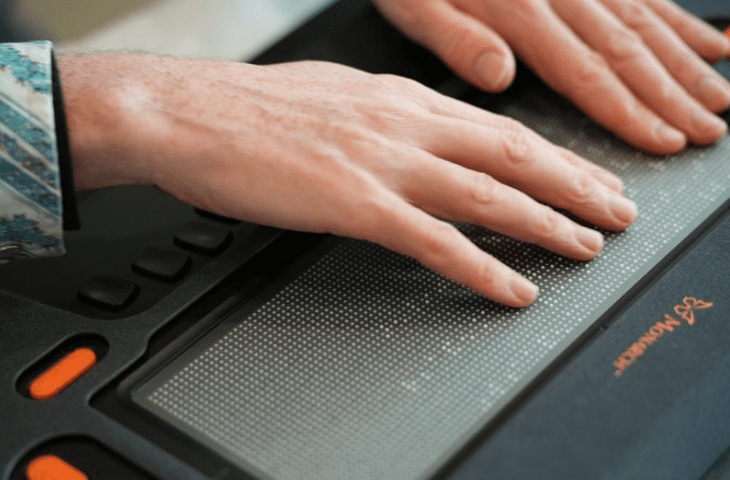 Two adult hands run their fingers across tactile braille on the Monarch multiline display.