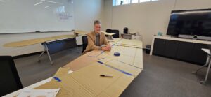 Darrick sits at a large desk wearing a tan suit. Across the desk are pieces of tape and cardboard.