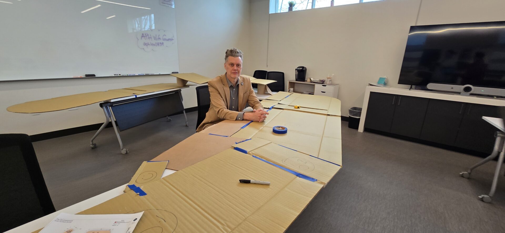 Darrick sits at a large desk wearing a tan suit. Across the desk are pieces of tape and cardboard.