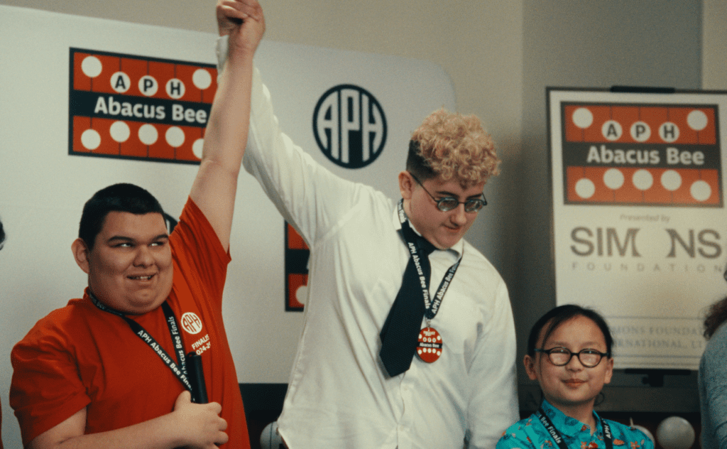 A smiling student in a red shirt and a student in a white shirt have their hands lifted together in the air. A third person smiles nearby. Signs in the background have the APH Abacus Bee, APH, and Simons Foundation logos.