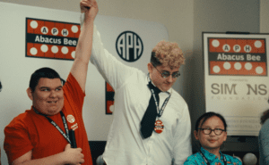 A smiling student in a red shirt and a student in a white shirt have their hands lifted together in the air. A third person smiles nearby. Signs in the background have the APH Abacus Bee, APH, and Simons Foundation logos.