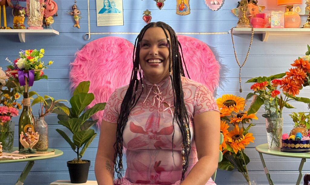 Precious sits in a room full of flowers and bright colors, wearing a soft pink tshirt. She smiles at the camera.