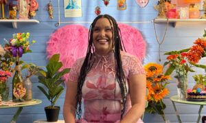 Precious sits in a room full of flowers and bright colors, wearing a soft pink tshirt. She smiles at the camera.