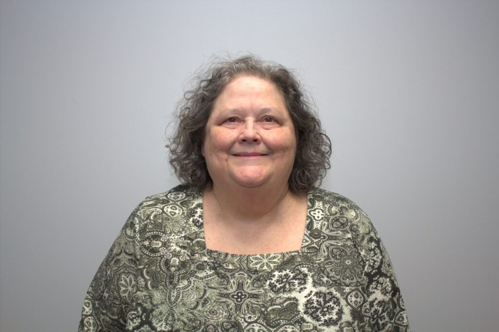 Jo Holt, wearing a floral green shirt, stands in front of a plain background smiling at the camera.