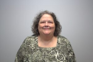 Jo Holt, wearing a floral green shirt, stands in front of a plain background smiling at the camera.